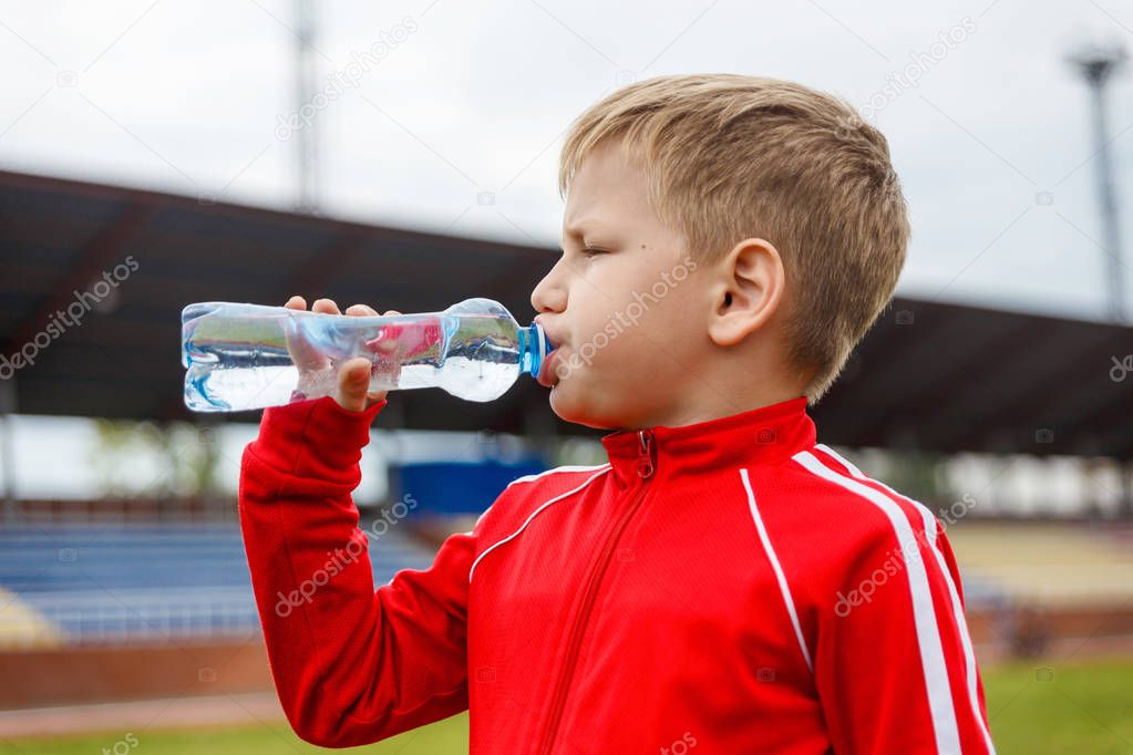 six-year-old boy in a red uniform drinking water from a small bottle at a sports stadium