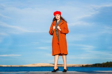 Turuncu ceket ve kırmızı bere genç kız mavi gökyüzüne karşı uzun duruyor ve kameraya benziyor