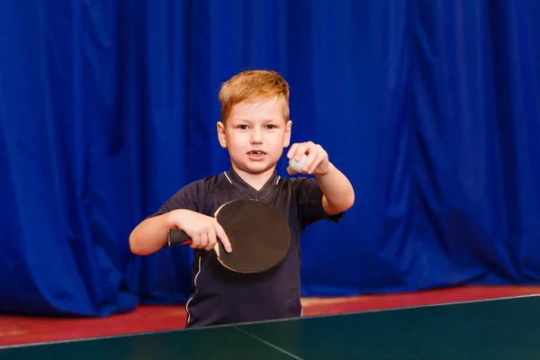 a child with a ball and a table tennis racket looks into the camera indoors