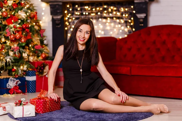 esmer kadın kısa siyah elbise katta oturan gülümsüyor. Noel arifesinde. Noel dekor kırmızı tarzı.