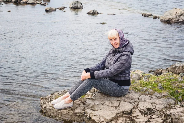 cute woman fifty years in a gray jacket and leggings sitting on a stone near the river in the spring