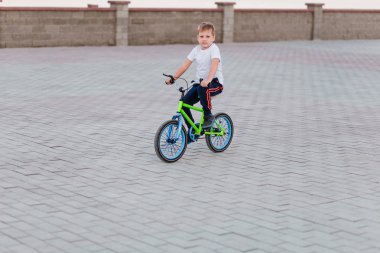 7 yaş çocuk beyaz bir t-shirt ile yaz aylarında yeşil Bisiklet Park şehirde kaldırım döşeme üzerinde gidiyor. Spor çocuk. Bisiklete binmek