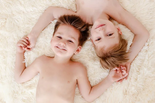 two smiling boys lie on their backs on a white fluffy blanket, top view