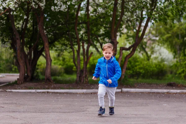 six-year-old Caucasian boy in a blue windbreaker and gray pants running in the Park in the spring