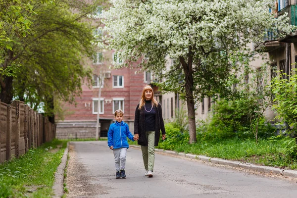 a woman of fifty walks by the hand with her seven-year-old grandson in the city quarter in the spring, blooming Apple trees in the background