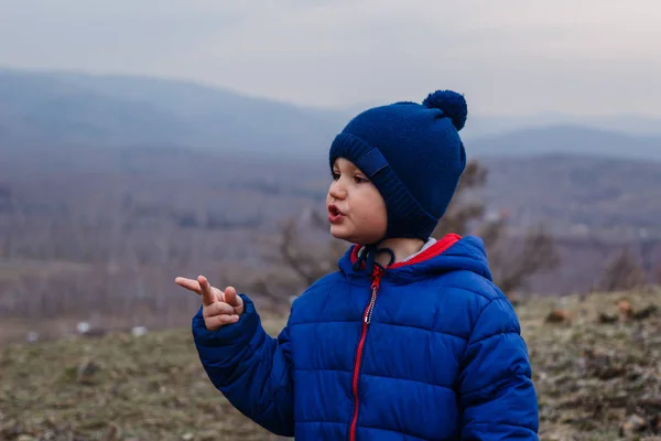 five-year-old boy in a cap with a pompom and a blue warm jacket on the background of the misty mountains in the spring