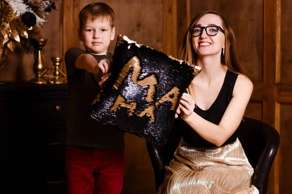 Smiling woman with glasses and a seven-year-old boy holding a Pillow with sequins. Stylish happy mother and son in the room. Mother's day.