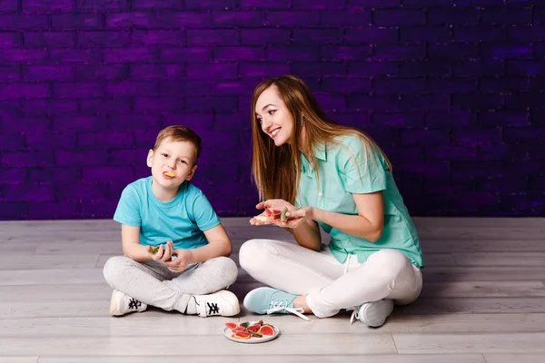 Ung långhårig mamma och sju-åriga son i skjortor och sneakers sitter på golvet och ha roligt. Vackra trettio år gammal kvinna och barn — Stockfoto