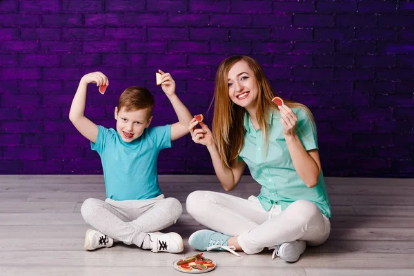 En ung kvinna som långhåriga och ett sju-åriga barn i skjortor och sneakers sitter på golvet och håll färgade skivor av marmelad i sina händer — Stockfoto