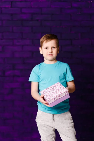 Menino de sete anos segurando uma caixa de presente no fundo roxo — Fotografia de Stock