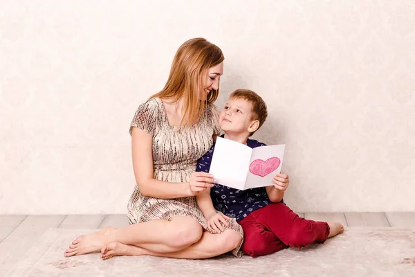 a young smiling mother looks at her seven-year-old son sitting on the carpet of the house. Greeting card for mom