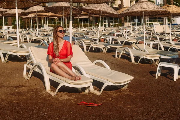 Vackra leende kaukasisk kvinna i röd transparent tunika och solglasögon liggande på en vit solsäng på stranden i ett hotell — Stockfoto