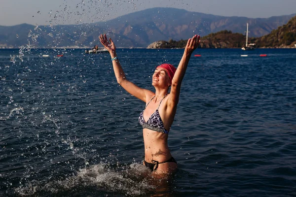 Donna snella trentenne in costume da bagno e una bandana spruzza acqua sul mare — Foto Stock