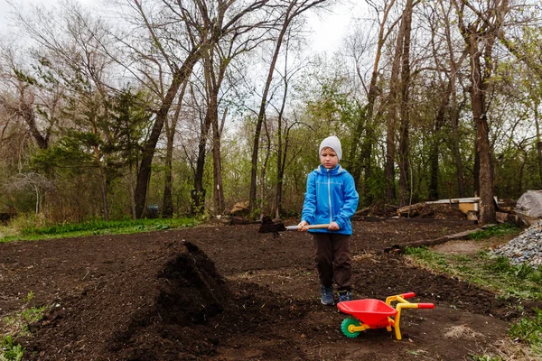 Семилетний мальчик в синей ветровке и шляпе бросает землю с саперной лопатой в телегу в саду — стоковое фото