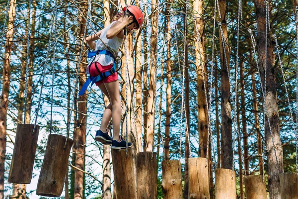 Ein zehnjähriges Mädchen in kurzen Hosen, T-Shirt und Schutzhelm läuft durch den Seilpark. auf Holzpfählen in luftiger Höhe. Extrem — Stockfoto