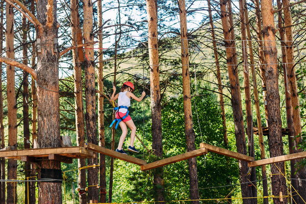 a ten-year-old girl in shorts, a t-shirt and a protective helmet passes through the rope Park.. Extreme sports on top.