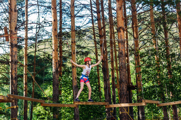 a brave sports ten-year-old girl in shorts, a t-shirt and a protective helmet is walking along the rope Park outdoors