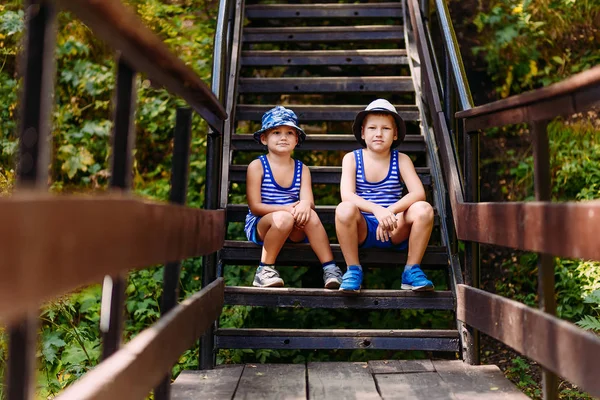 Два маленьких мальчика в футболках, кроссовках и шляпах сидят на ступеньках летом — стоковое фото