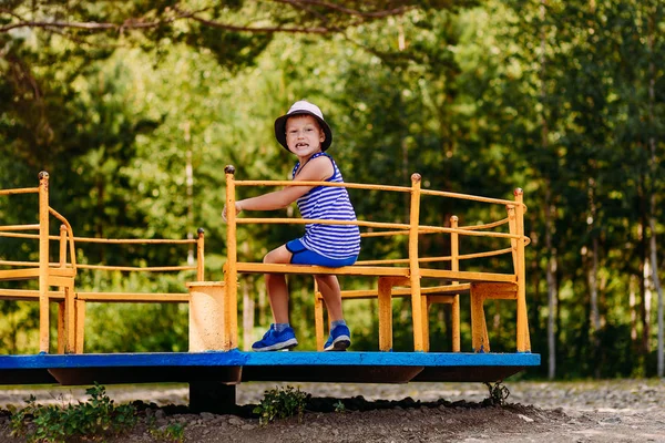 Garçon gai de sept ans dans un t-shirt rayé et chapeau assis sur un carrousel jaune pour enfants — Photo