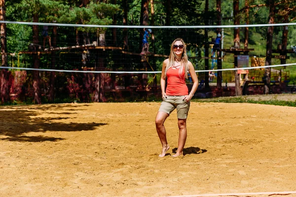Спортивная улыбающаяся загорелая женщина в солнцезащитных очках стоит на желтом песке на пляже — стоковое фото