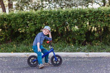 Şapkalı dört yaşında gülümseyen bir çocuk mavi koşu bisikletinin üzerinde oturuyor. Pedalsız bisiklet