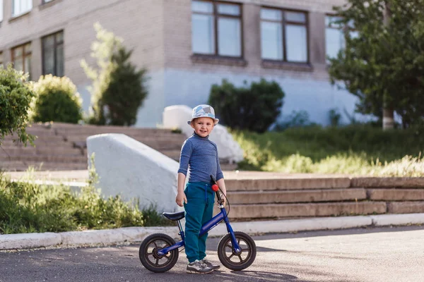 A four-year-old smiling boy in a hat sits on a blue running bike. Bicycle without pedals — Stock Photo, Image