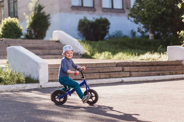 Um menino alegre de quatro anos de idade em um chapéu e tênis monta uma bicicleta de corrida e empurra seus pés fora do asfalto . — Fotografia de Stock