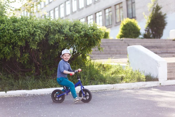 A four-year-old cheerful boy in a hat and sneakers rides a running bike and pushes his feet off the asphalt. — Stock Photo, Image