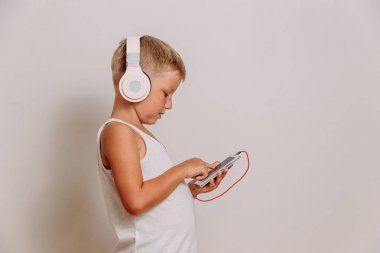 yedi yaşındaki bir çocuk beyaz arka plan üzerinde büyük tam boyutlu kulaklıklar aracılığıyla müzik dinler. Çocuk akıllı telefon kullanıyor..