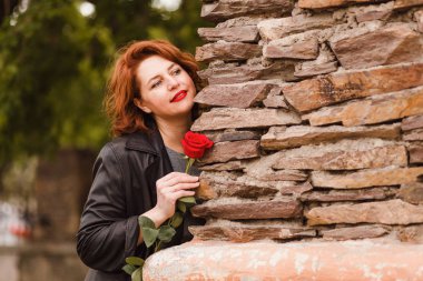 kırmızı bir gül tutan deri siyah pelerin kırmızı dudakları ile gülümseyen orta yaşlı kadın