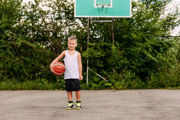 Маленький баскетболист в белой футболке и черных шортах стоит с мячом на баскетбольной площадке летом — стоковое фото