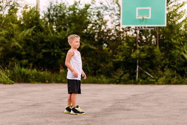 Семилетний мальчик в баскетбольной форме летом тренируется на открытой баскетбольной площадке. Дети и спорт, мальчик в баскетбольных кроссовках — стоковое фото
