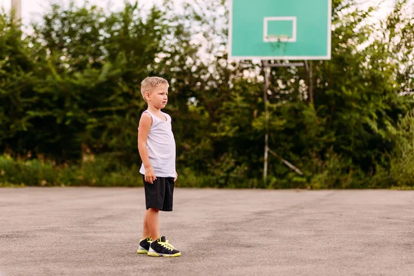 Семилетний мальчик в баскетбольной форме летом тренируется на открытой баскетбольной площадке. Дети и спорт, мальчик в баскетбольных кроссовках и белой футболке — стоковое фото