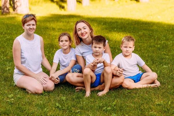 Mujeres sonrientes y niños se sientan en la hierba verde. Gran familia feliz, dos madres y tres niños con camisetas blancas. Verano — Foto de Stock