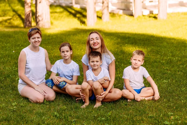 Happy women and children sit on the green grass and look at the camera. Big happy family, two mothers and three children in white t-shirts and shorts. — Stok fotoğraf