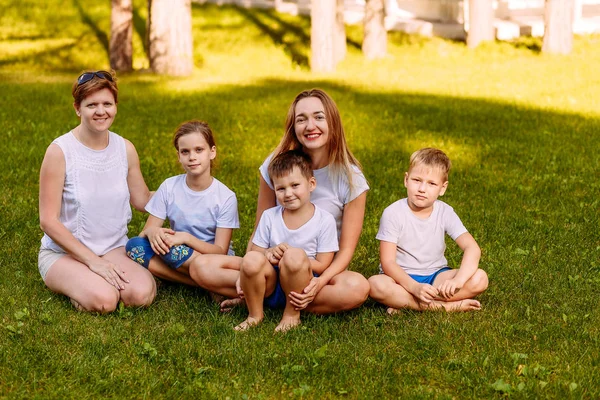 Szczęśliwe młode kobiety i dzieci siedzą na zielonej trawie i patrzą na kamerę. Duża szczęśliwa Rodzina na wakacjach, dwie matki i troje dzieci w białych koszulkach i szortach. Letnich — Zdjęcie stockowe