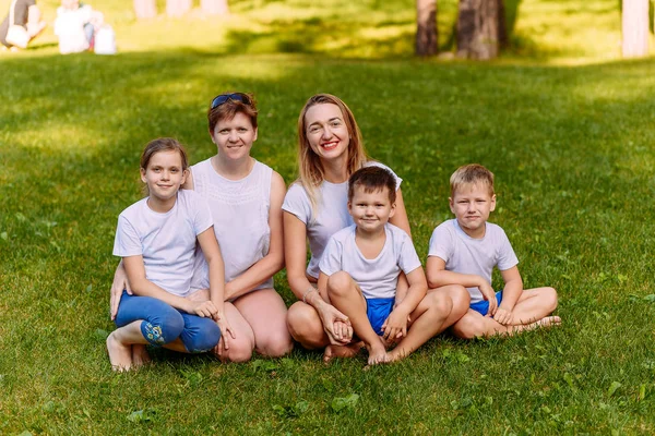Dos madres jóvenes felices y tres niños con camisetas blancas se sientan en un césped verde en el verano. Una gran familia caucásica de vacaciones. Mujeres abrazan a los niños . — Foto de Stock