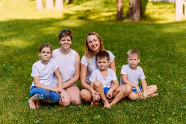 Två lyckliga unga mödrar och tre barn i vita t-shirts sitter på en grön gräsmatta på sommaren. Stor kaukasisk familj på semester — Stockfoto