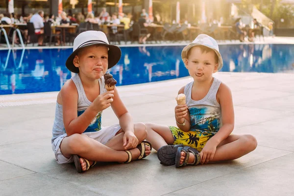 two pre-school boys in hats sit by the hotel's outdoor pool and eat ice cream in a waffle cone . Kids and ice cream. Lips in ice cream