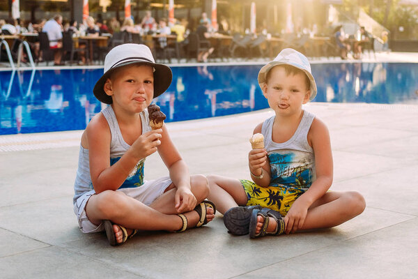 two pre-school boys in hats sit by the hotel's outdoor pool and eat ice cream in a waffle cone . Kids and ice cream. Lips in ice cream