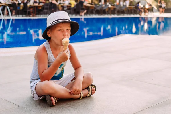 Семилетний мальчик в шляпе, сидит у открытого бассейна и ест мороженое в вафельном конусе — стоковое фото