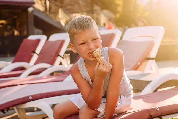 Семилетний блондин ест мороженое в вафельном рожке у бассейна на закате солнца при свете солнца. — стоковое фото