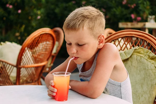 Sedmiletý blonďatý chlapec, který pije pomerančový nealkoholický koktejl z trubice v letní restauraci — Stock fotografie