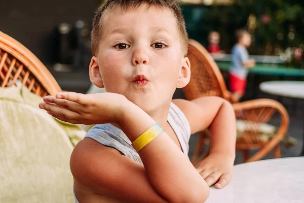 Пятилетний белый мальчик гримасы за столом в летнем кафе. Дети и летние каникулы. Hotel bracelet on hand — стоковое фото