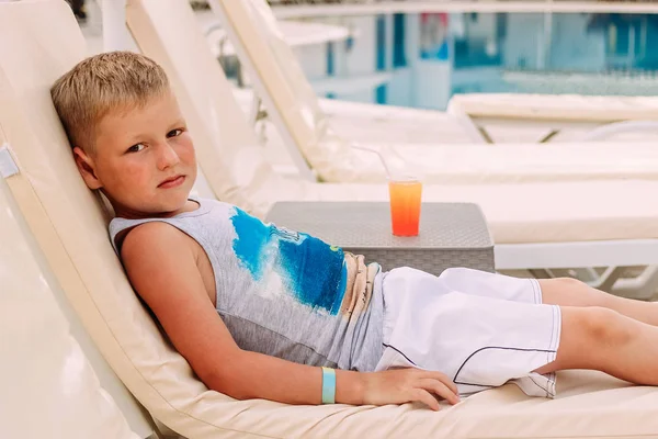 Un lindo chico rubio caucásico de siete años yace junto a la piscina al aire libre . —  Fotos de Stock