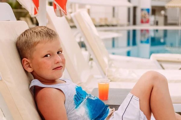 Um menino bonito loiro caucasiano de sete anos está deitado à beira da piscina exterior. cocktail de fruta laranja  . — Fotografia de Stock