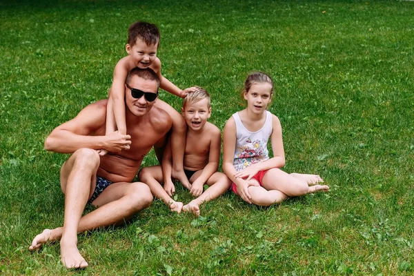 Opalony młody ojciec w ciemnych okularach i trzy wesołe dzieci siedzą na trawie w lecie. Dzień Ojca, ojciec z wieloma dziećmi — Zdjęcie stockowe