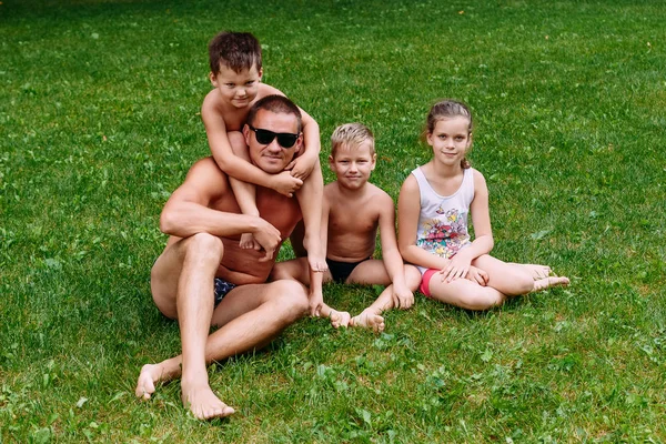 Garvade ung man i mörka glasögon och tre barn som sitter på gräset på sommaren. Fars dag — Stockfoto