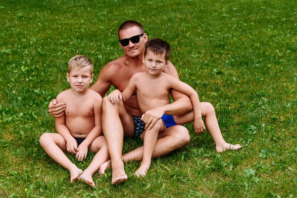 Счастливый молодой отец обнимает двух своих сыновей летом на зеленой лужайке. Мужчина и дети — стоковое фото