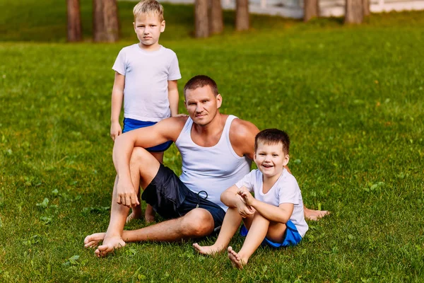 Отец и двое детей летом сидят на зеленой лужайке. Мужчина и два маленьких сына дошкольника летом — стоковое фото